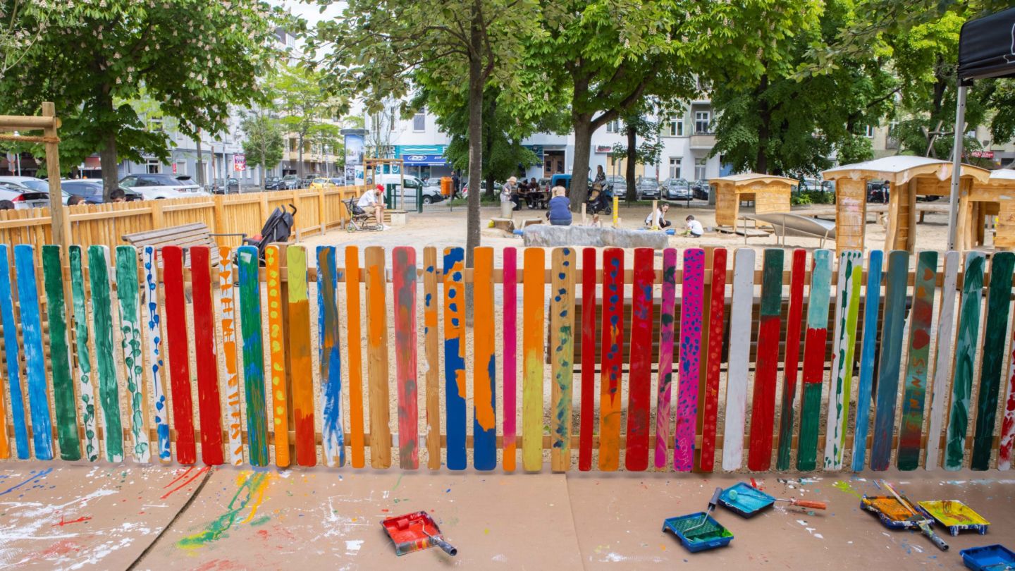 Bunt gestrichener Zaun, mit Farbkästen und Rollen im Vordergrund